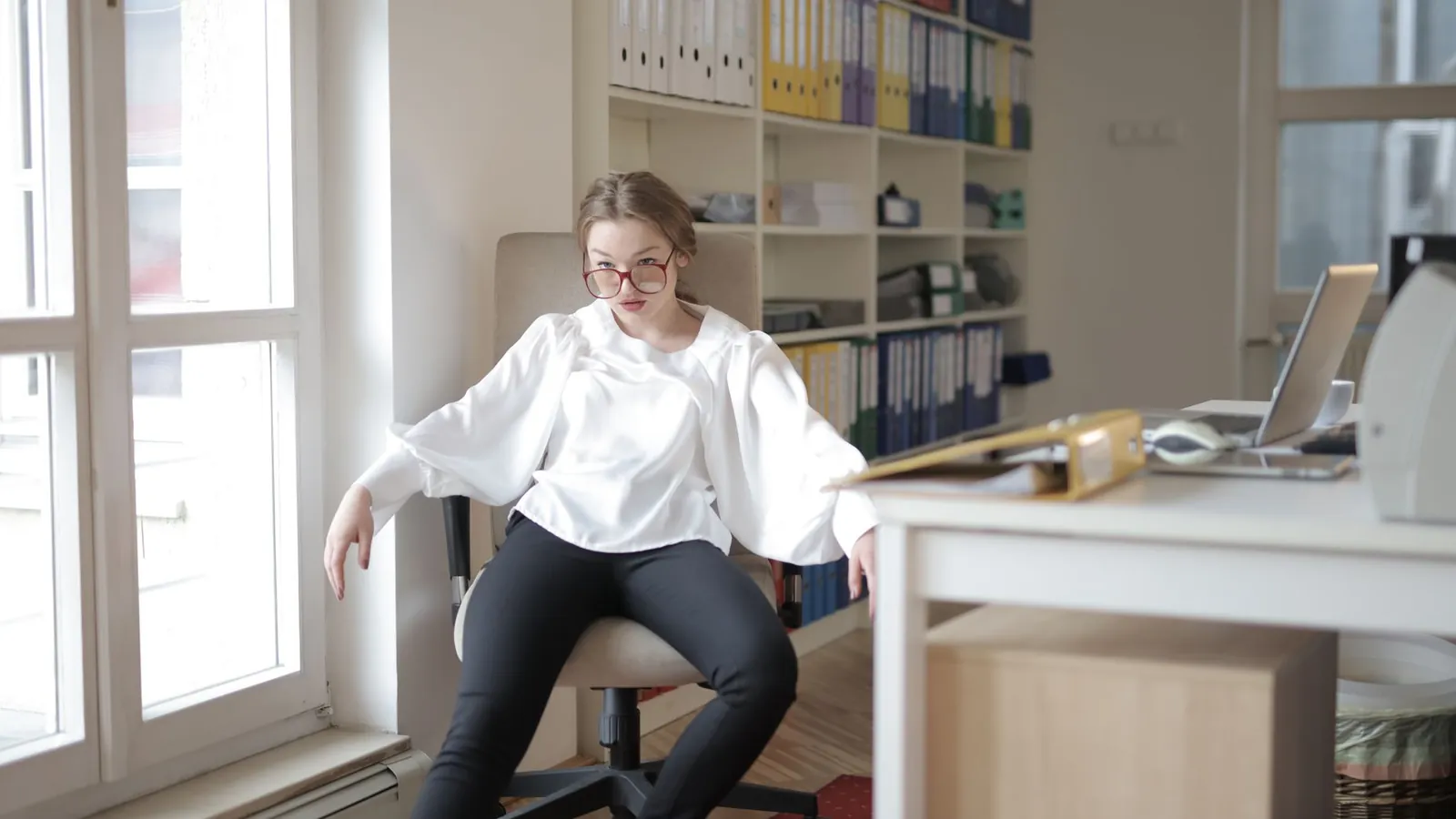 A person sitting in a relaxed slouch on an office chair by a window