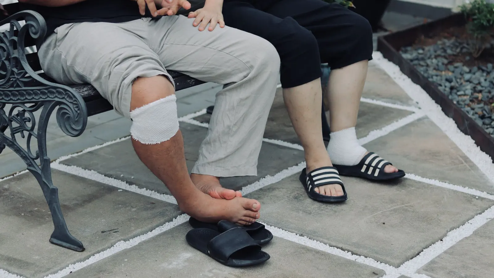 A person with a bandaged knee resting on a bench beside a walking stick